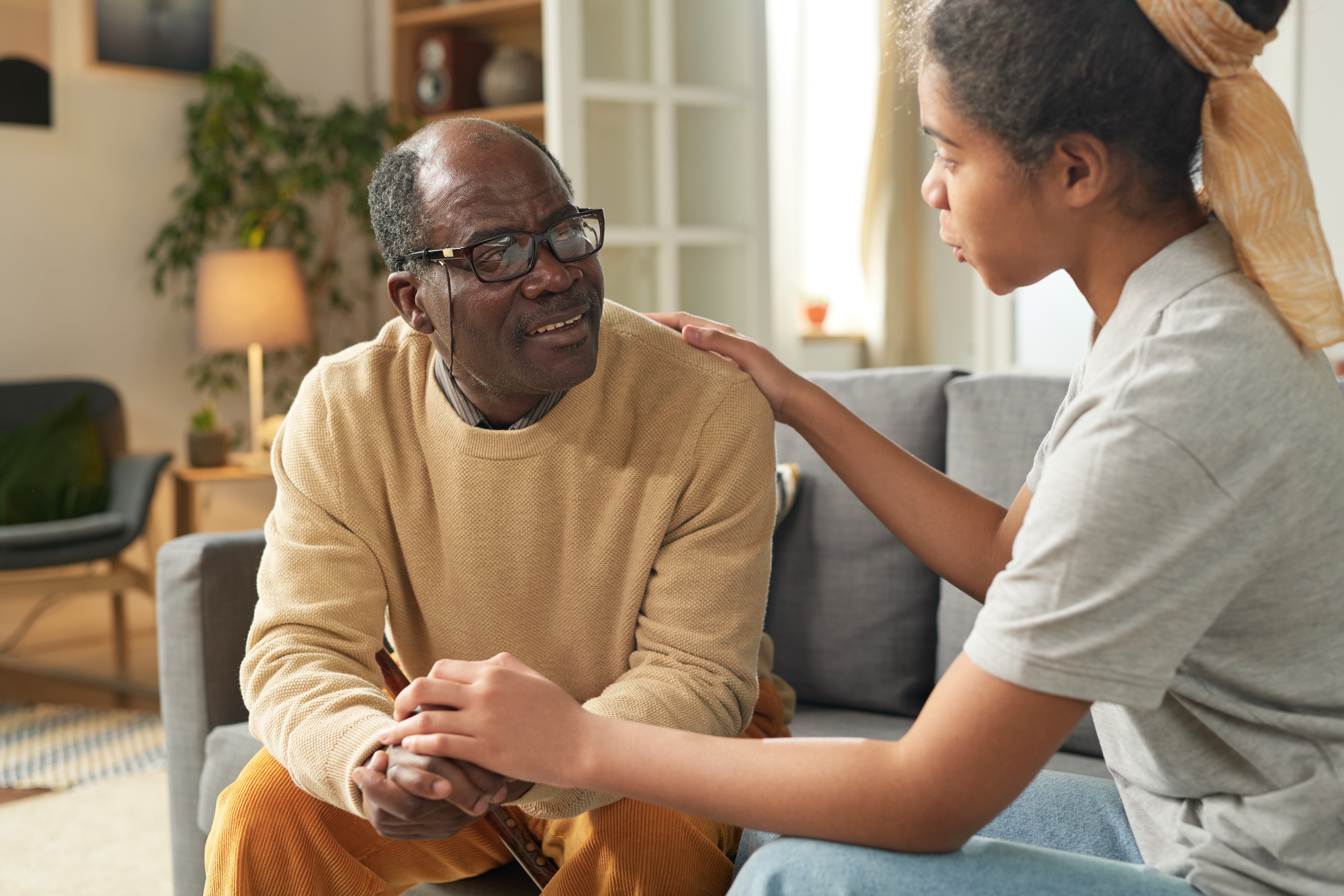Elderly man sitting in living room being comforted by young African American girl holding his hand expressing care and empathy